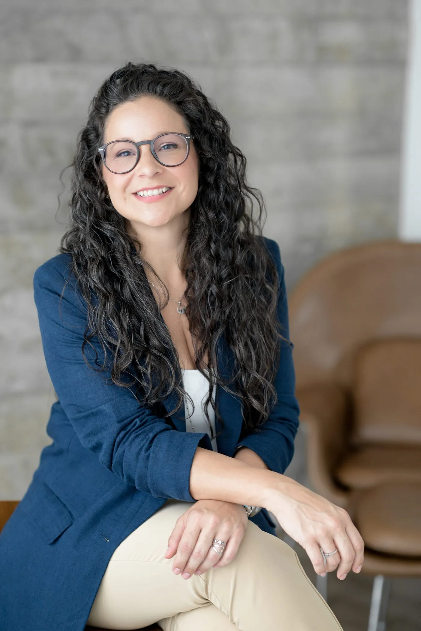Retrato de Camila Gracioli sorrindo, de óculos e blazer azul, sentada com as mãos sobre o joelho.