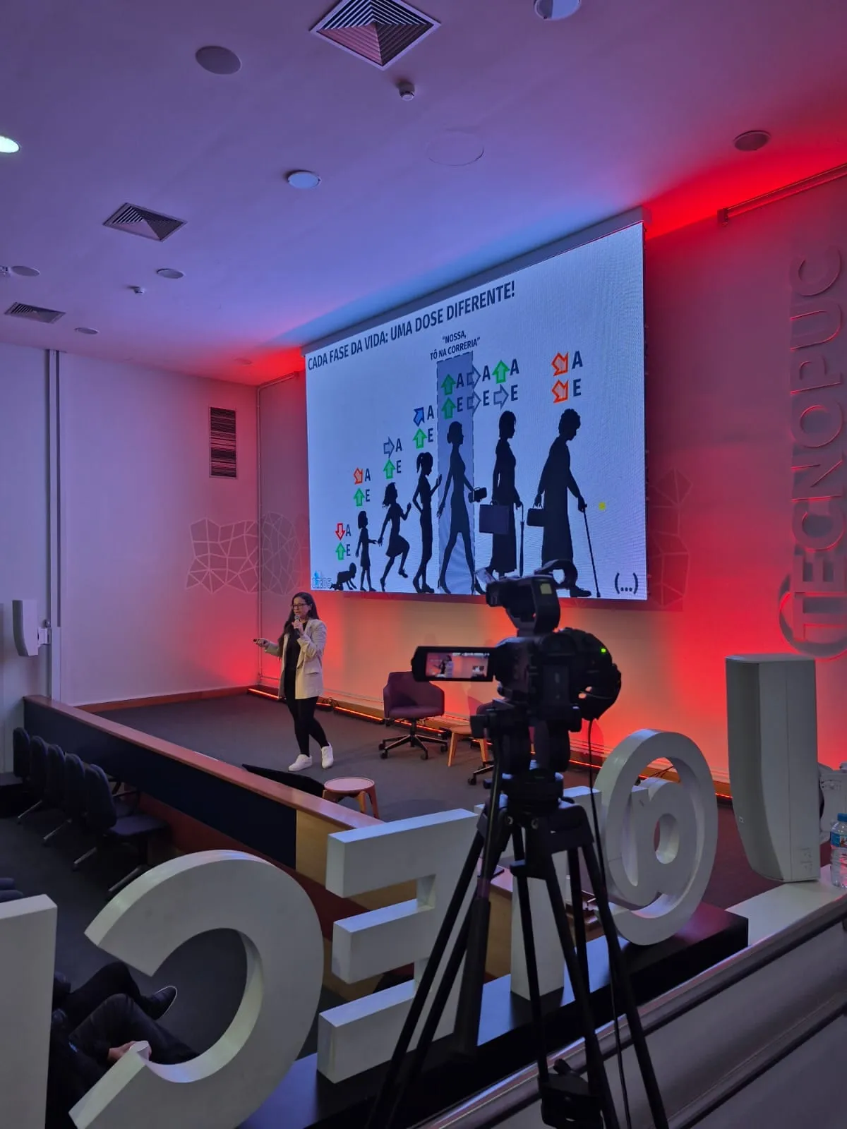 Camila Gracioli no palco de um auditório, com uma câmera de filmagem em primeiro plano.